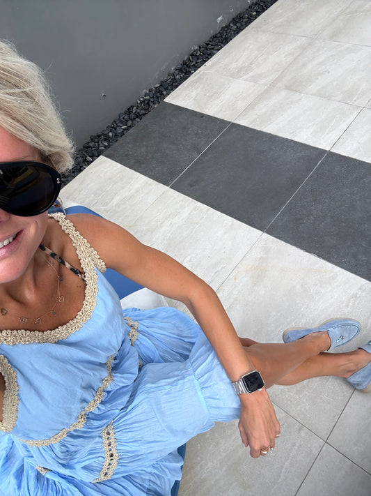 A woman wearing a light blue ruffled dress with beige crochet trim, light blue loafers, and black sunglasses, sitting on a blue and white striped lounge chair on a tiled patio.