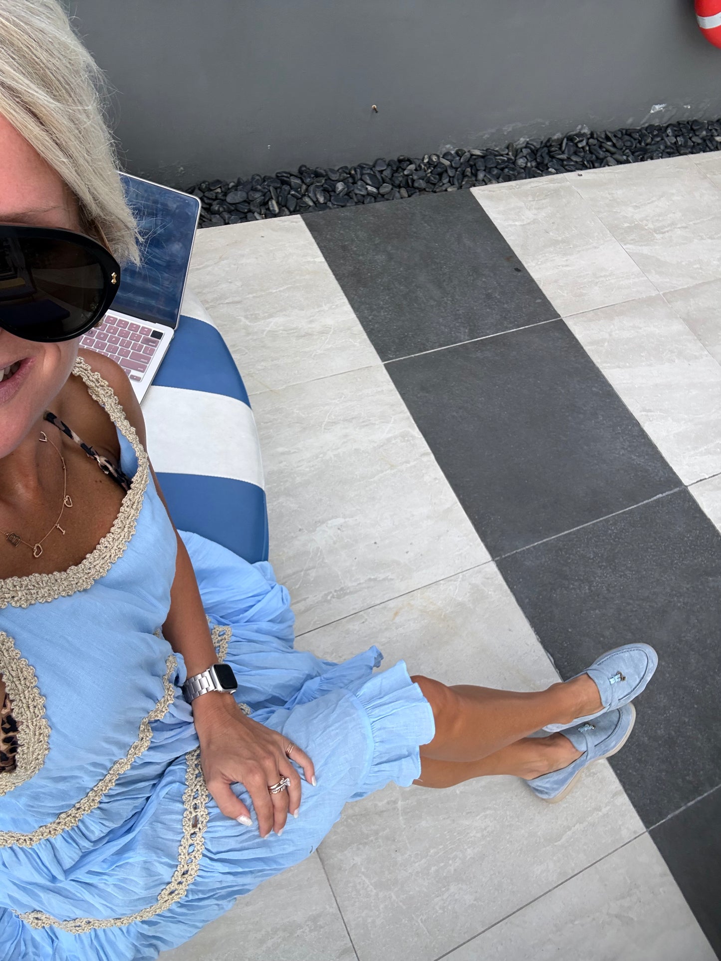 A woman wearing a light blue ruffled dress with beige crochet trim, light blue loafers, and black sunglasses, sitting on a blue and white striped lounge chair on a tiled patio.